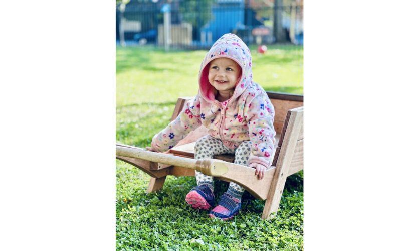 Preschool girl in hooded coat sitting in wooden wheelbarrow